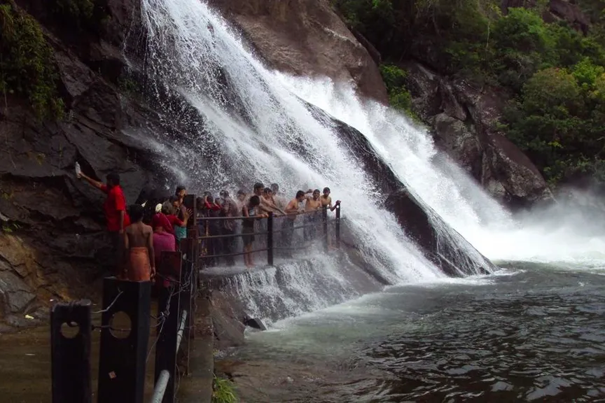 Visitors enjoying the cascading Ayyanar Falls in Tamilnadu, standing on a wet rock platform beside the flowing water.