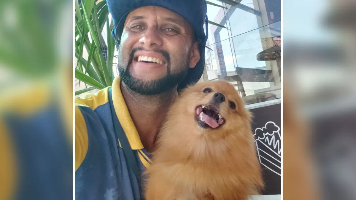 Smiling mailman wearing a blue hat poses happily with a fluffy, joyful brown dog in his arms.